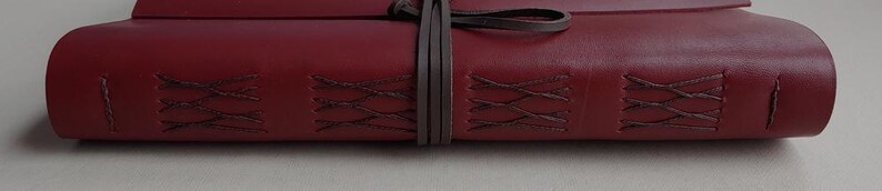May include: A burgundy leather journal with decorative stitching and a wraparound leather tie. The journal's cover features a textured surface and a dark brown tie to keep it closed. The stitching is a darker shade of brown.