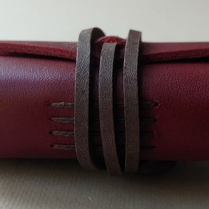 May include: A burgundy leather journal with brown leather straps wrapped around it. The journal has stitching details on the edges.