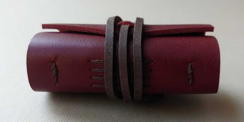 May include: A burgundy leather journal with brown leather straps wrapped around it. The journal has stitching details on the edges.