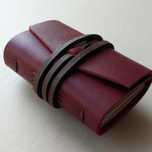 May include: A small burgundy leather journal with a brown leather strap closure. The journal is closed and lying on a white surface.