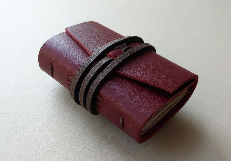May include: A small burgundy leather journal with a brown leather strap closure. The journal is closed and lying on a white surface.