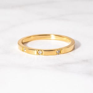 Può includere: Un anello a fascia dorato con tre piccole pietre preziose trasparenti, equidistanti sulla fascia. L'anello ha un profilo piatto e rettangolare e una finitura lucida. L'anello è posto su uno sfondo di marmo bianco.