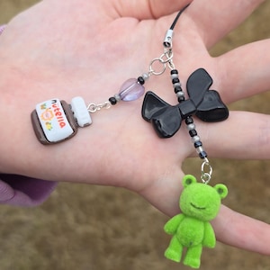 May include: A handmade charm featuring a miniature Nutella jar, a black bow, and a green fuzzy frog. The charm is attached to a black cord with silver-colored beads and a purple heart-shaped bead. The Nutella jar has the brand name printed on it.