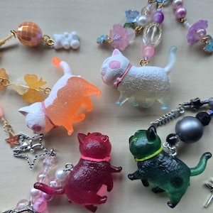 May include: A collection of colorful cat-shaped charms with beaded accents. The charms are in shades of orange, white, red, and green, each with a collar. The beaded strands include pearls, stars, and floral elements, attached to a blue clasp.