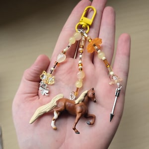 May include: A brown pegasus keychain with a yellow clasp, adorned with beaded strands in shades of yellow, orange, and brown. The keychain features a small silver arrow charm and a butterfly charm.
