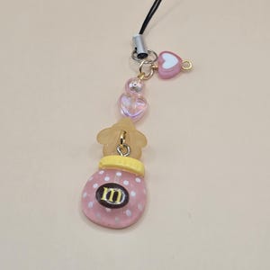 May include: A pink and white beaded charm with a candy jar design. The charm features a pink jar with white polka dots, a yellow lid, and the letter "M" in the center. The charm also includes clear heart-shaped beads and a pink heart charm.