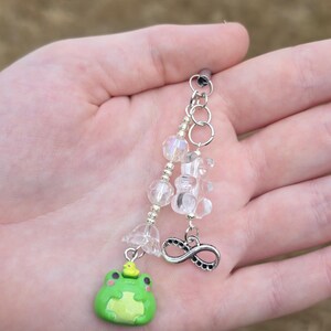 May include: A handmade frog charm with a silver chain, clear beads, and an infinity symbol. The frog is green with a yellow crown and a light green belly. The charm is held in a person's hand.