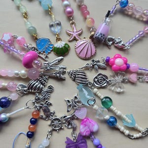 May include: A collection of beaded phone charms in various colors. Charms include seashells, starfish, flowers, and a boba tea design. Beads are in shades of pink, purple, blue, green, and white. Each charm has a silver-tone clasp.