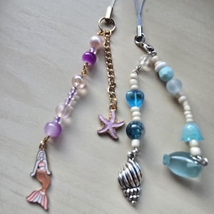May include: Three beaded phone charms with ocean-themed charms. One charm features a mermaid, purple beads, and a gold chain. Another has a starfish, and the third has a seashell and a blue glass fish.