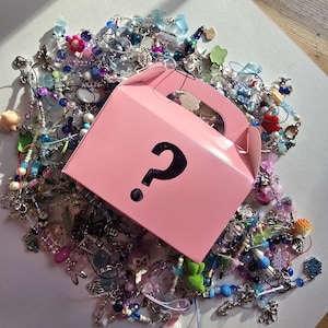 May include: A pink box with a black question mark sits atop a pile of colorful beads, charms, and trinkets. The beads are in shades of blue, purple, pink, and white. The charms include flowers, animals, and other decorative elements. The box suggests a mystery item.