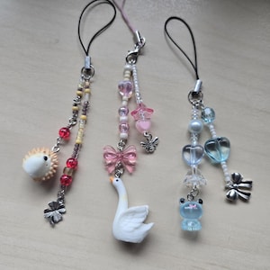 May include: Three decorative phone charms with various designs. One features a hedgehog, another a swan with a pink bow, and the third a blue frog. Each charm has a beaded strand and a silver-tone clasp.