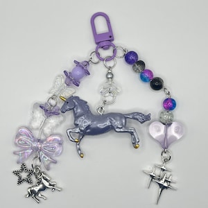 May include: A purple unicorn charm with a silver star, a silver unicorn, and a purple heart charm. The charm is attached to a silver chain with purple, blue, and clear beads.