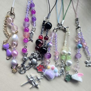 May include: A collection of beaded phone charms in various colors, including purple, pink, and white. Each charm features unique decorative elements such as cat and dog charms, paw prints, and other small pendants. The charms are attached to white cords.
