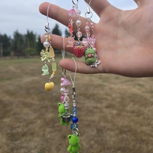 May include: A collection of handmade phone charms with various colorful beads and charms. The charms include a yellow heart, green frogs, pink strawberries, and butterflies. The charms are attached to a white string and are held up against a blurred outdoor background.