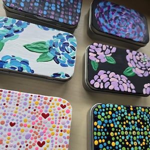 May include: Six rectangular metal tins featuring hand-painted designs. The top left has blue and purple dots on black, the top right has purple and blue swirls, the middle left has blue flowers on white, the middle right has pink flowers on black, the bottom left has colorful dots and hearts on white, and the bottom right has a green, blue, and yellow dot pattern on black.