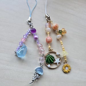 May include: Two beaded phone charms with ocean-themed pendants. One charm features a blue jellyfish and a blue gem, while the other has a seashell, starfish, and sunflower charm. The beads are in shades of pink, purple, and orange.