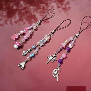 May include: Three beaded phone charms with black loops. One charm features a pink butterfly and mushroom, another has a mermaid tail charm, and the third has a star and moon charm. The beads are pink, purple, and clear.