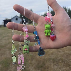 May include: A collection of colorful beaded charms featuring frog and strawberry pendants. The charms include pink, green, and blue beads, with small charms like butterflies, bows, and hearts. Each charm is attached to a white string.