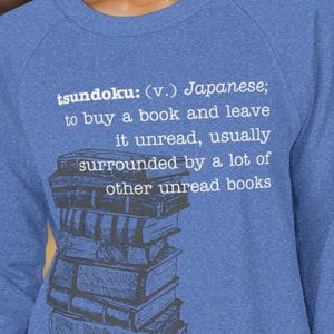 May include: Blue sweatshirt with the definition of "tsundoku" printed on it. The text describes the Japanese word for buying books and leaving them unread, usually surrounded by other unread books. A stack of books is illustrated below the text.