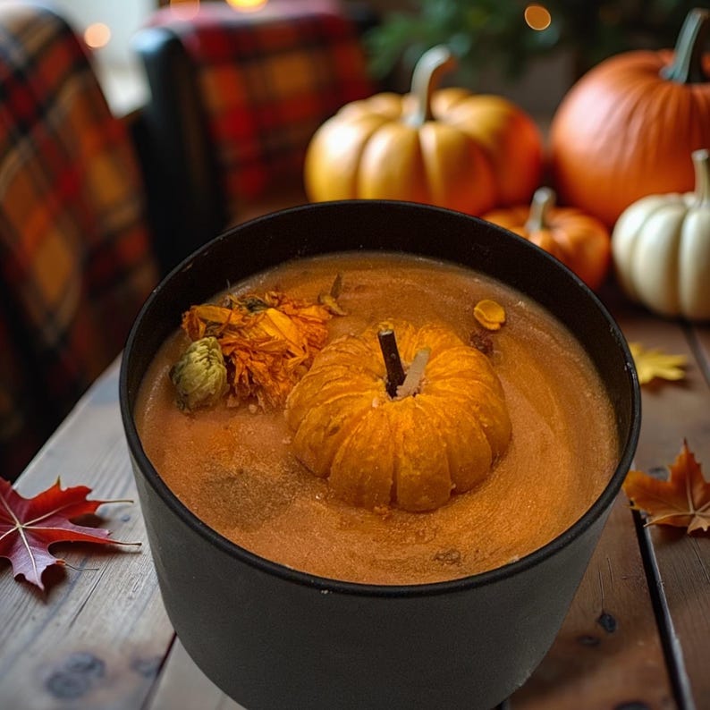May include: A black candle jar with a pumpkin-shaped orange candle inside. The candle is surrounded by a brown liquid and a dried flower. The jar is sitting on a wooden table with pumpkins in the background.