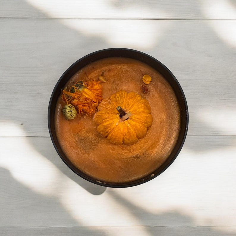 May include: A black bowl filled with a pumpkin spice candle. The candle has a small orange pumpkin floating in the center and dried orange flowers around the pumpkin.