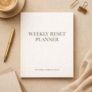 Pode incluir: Um Weekly Reset Planner creme com detalhes dourados. A capa apresenta o texto "WEEKLY RESET PLANNER" e "THE STRUCTURED WOMAN" em uma fonte marrom escuro. Uma caneta dourada, clipes de papel e uma xícara de café também são visíveis.