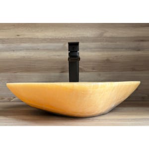May include: A light orange, oval-shaped vessel sink with a black faucet. The sink is made of a translucent material, and the faucet is a modern, minimalist design. The sink is set against a light brown wooden background.