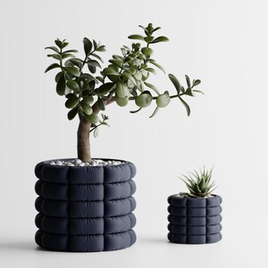 May include: Two cylindrical planters with a dark navy blue, quilted, segmented exterior texture. The larger planter holds a jade plant with green leaves, while the smaller planter contains a small succulent. Both are topped with white pebbles.