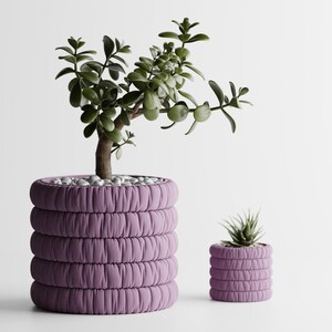 May include: Two cylindrical planters in a soft lavender color, each featuring a distinct horizontal ribbed texture that resembles gathered fabric. The larger planter holds a green succulent plant, while the smaller one contains a tiny succulent.