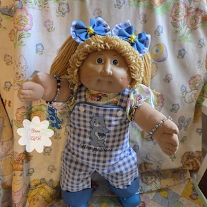 Puede incluir: Una muñeca con cabello de lana rubia peinado en coletas con lazos de cuadros vichy azules y blancos. La muñeca viste un peto de cuadros vichy azules y blancos con un gráfico de un oso azul y el texto Grumpy Bear, sobre una camisa estampada.
