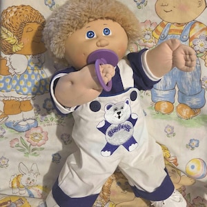 Puede incluir: Muñeca vintage Cabbage Patch Kids con un atuendo blanco y azul marino con un motivo de oso. La muñeca tiene cabello rubio, ojos azules y un chupete morado. La muñeca está sobre una manta estampada.