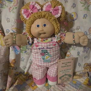 Puede incluir: Una muñeca Cabbage Patch Kids con cabello de lana rubia peinado en dos coletas con lazos de cuadros rosas y blancos. La muñeca viste un peto de cuadros rosas y blancos con un gráfico y texto de Wish Bear, e incluye un certificado de nacimiento.