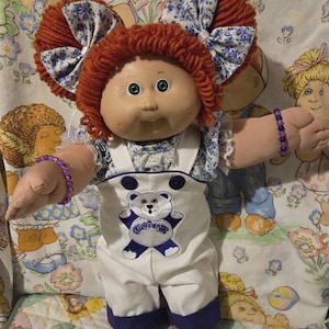 Puede incluir: Muñeca vintage Cabbage Patch Kids con pelo de hilo rojo y ojos verdes. La muñeca viste un atuendo blanco con un diseño de oso de peluche azul y detalles azules a juego. Tiene un lazo con estampado floral y pulseras moradas.