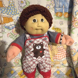 May include: A doll with brown yarn hair, light skin, and dimples, wearing a gray shirt with red trim and white plaid overalls featuring a brown teddy bear graphic with a red heart. A small beaded bracelet is on the doll's left wrist.