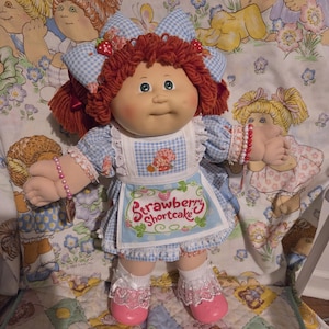Puede incluir: Muñeca vintage de Strawberry Shortcake con cabello rojo de hilo, ojos azules y mejillas rosadas. Viste un vestido de cuadros azul y blanco, un gorro a juego y un delantal de Strawberry Shortcake. Sus zapatos rosas y calcetines de encaje blanco completan el look.