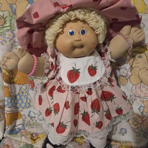 Puede incluir: Muñeca vintage de Cabbage Patch Kids con un atuendo rosa con temática de fresas. La muñeca tiene cabello rubio rizado, ojos azules y un gorro rosa. El vestido y el gorro están adornados con fresas rojas. La muñeca lleva zapatos negros y un babero blanco.