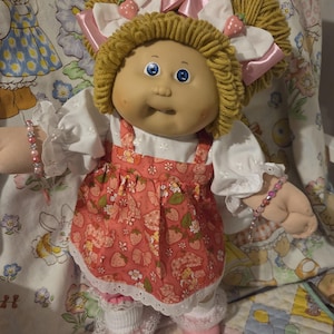 May include: A doll with yarn hair styled in two pigtails, blue eyes, and dimpled cheeks. The doll wears a white blouse with eyelet lace sleeves, a pink strawberry-patterned pinafore dress, white lace-trimmed socks, and pink shoes with beaded bracelets.