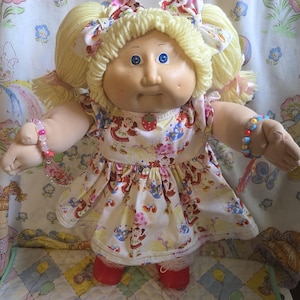 Könnte beinhalten: Eine Puppe mit blasser Haut, blauen Augen und Sommersprossen, mit gelbem Wollhaar, das zu zwei Zöpfen mit Blumen-Schleifen gebunden ist. Die Puppe trägt ein weißes Kleid mit buntem Motivdruck, einen Erdbeer-Anhänger, rote Schuhe und Perlenarmbänder an den Handgelenken.