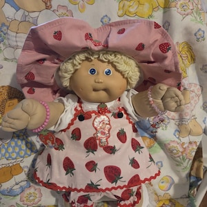 Puede incluir: Muñeca vintage Cabbage Patch Kids con un atuendo rosa con temática de fresas y un gorro a juego. La muñeca tiene cabello rubio rizado y ojos azules. El atuendo incluye un vestido, pantalones y zapatos, todos adornados con patrones de fresas.