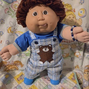 Peut inclure: Une poupée aux cheveux en laine brune et aux yeux marron, vêtue d'un t-shirt bleu sous une salopette à carreaux bleus et blancs ornée d'un motif d'ours en peluche marron. La poupée porte un bracelet de perles et des chaussures bleues.