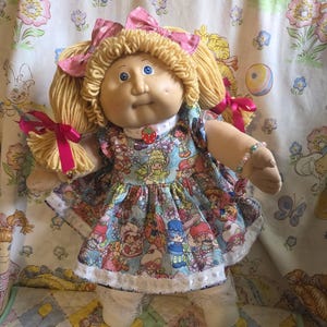 May include: A doll with blonde yarn hair styled in pigtails with pink bows, blue eyes, and freckles. The doll wears a dress featuring a colorful Strawberry Shortcake print, a small strawberry charm at the neckline, white lace-trimmed socks, and white shoes.