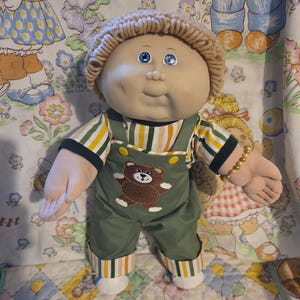 Op de afbeelding: Een vintage Cabbage Patch Kid pop met blauwe ogen, met een beige gebreide muts, een gestreept shirt met groene, gele en witte strepen en een groene overall met een bruine berenapplicatie. De pop heeft een gouden kralenarmband.