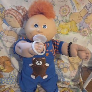 Pode incluir: Uma boneca Cabbage Patch Kid vintage com cabelo laranja e olhos azuis. A boneca usa um macacão azul com uma aplicação de urso marrom e uma camisa listrada laranja e azul. Tem uma chupeta na boca e uma pulseira de contas pretas.