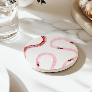 Könnte beinhalten: Ein runder weißer Keramikuntersetzer mit einem verspielten Design aus rosa und roten Rennstrecken und kleinen roten Rennwagen. Der Untersetzer liegt auf einer weißen Marmoroberfläche.