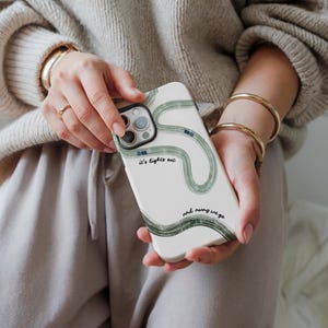 Puede incluir: Una funda de teléfono blanca con un diseño de carretera sinuosa verde y el texto "It's lights out and away we go". La funda la sostiene una persona que lleva un suéter beige y pulseras doradas.