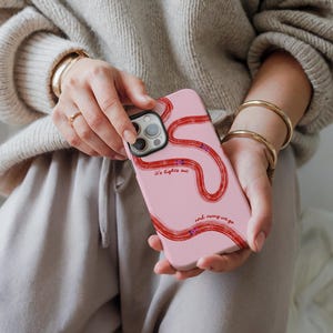 May include: Pink phone case with a red, wavy design and the text "It's lights out and away we go." The case is held by a person wearing a beige sweater and gold bracelets.