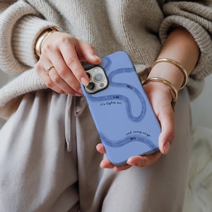 May include: A periwinkle blue phone case with a wavy, abstract design in a darker blue. The case features the text "It's lights me" and "and round we go". The phone case is held in a person's hands.