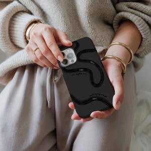 May include: Black phone case with a wavy, road-like design in black. The case has the text "It's lights me" and "and away we go" in a handwritten style. The case is held in a person's hands.