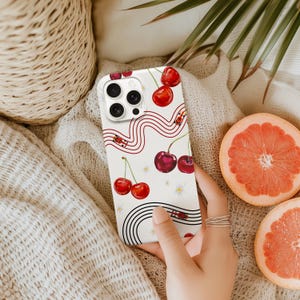 Puede incluir: Funda de teléfono blanca con diseño de cerezas. La funda presenta cerezas rojas, líneas onduladas rojas e ilustraciones de pequeñas mariquitas. La funda del teléfono se muestra sobre una superficie suave y texturizada con media toronja en el fondo.