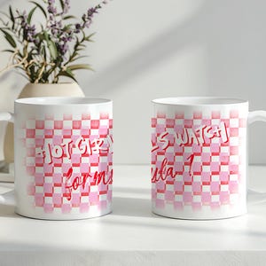 Puede incluir: Dos tazas de cerámica blanca con un patrón de cuadros rosas y rojos. Una taza tiene el texto "Hot Girl Formula" y la otra dice "Let's Watch Formula 1". Un jarrón con flores moradas está en el fondo.